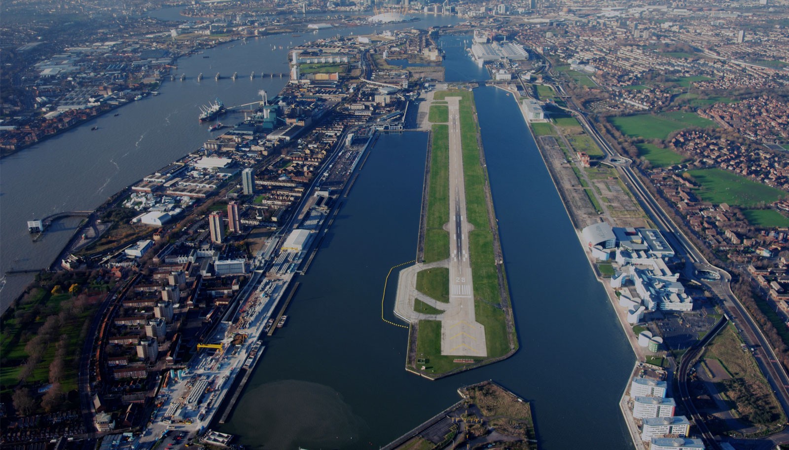 Aerial view of London City Airport with its runway surrounded by Royal Docks and Thames River, showcasing East London's urban landscape