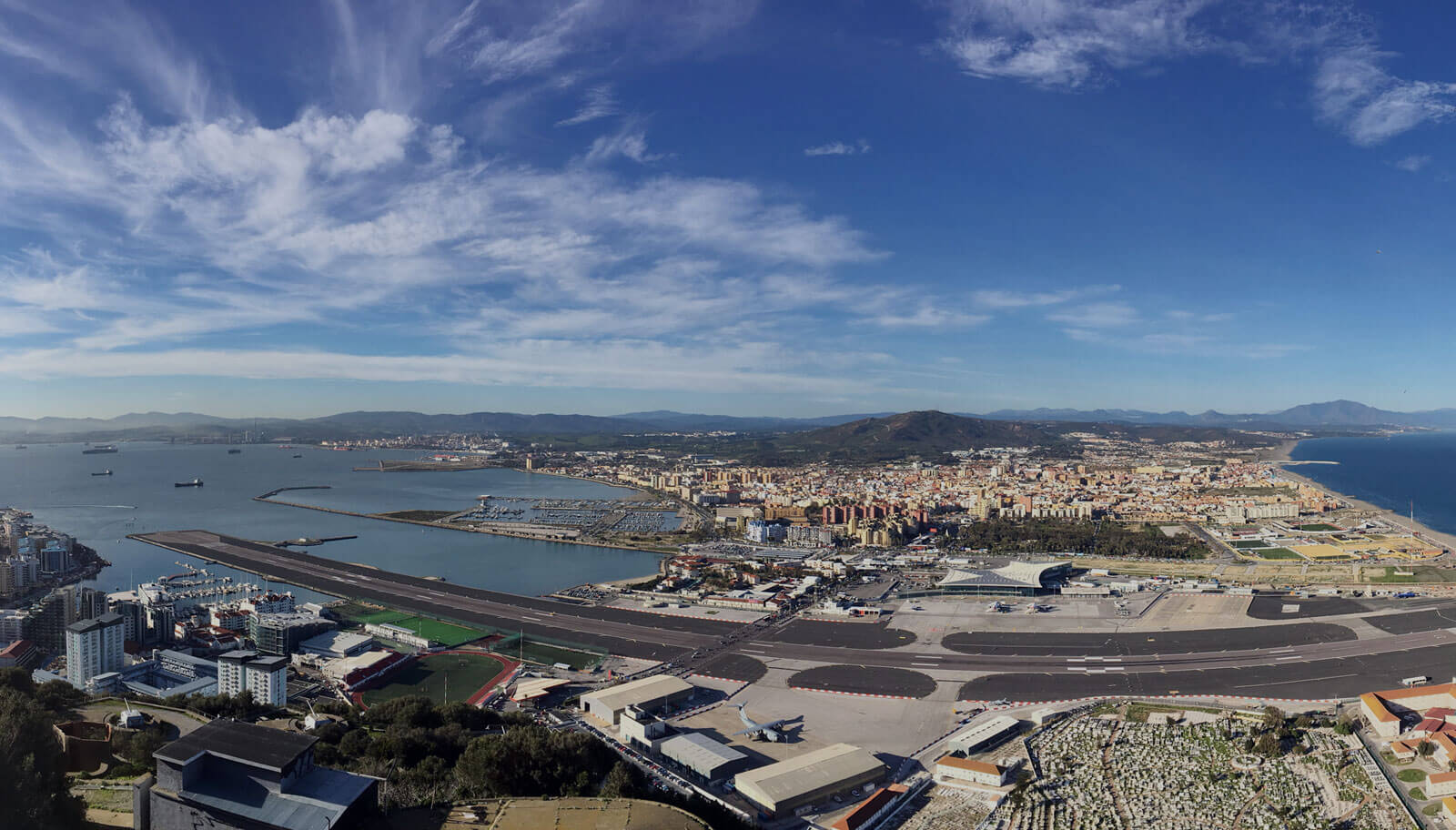 Panoramic aerial view of Gibraltar Airport with runway extending into the bay, cityscape, Mediterranean coastline, and mountains in background